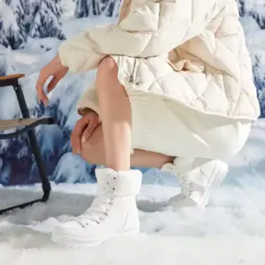 A woman in a light coat, skirt, and white fur-lined Wholesale Customize logo Leather Sports Ankle Sneakers Boots for Women Snow Waterproof Outdoor Warm Boot squats on snow by a stool with mountains, inspiring OEM ODM winter collections.