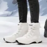 A person in white OEM ODM Source Factory Winter Waterproof Anti slip Warm Snow Boots Women's Solid PU Cotton Shoes and black jeans stands on snow before mountains, showcasing the boots' quality.