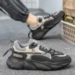 A person in dark joggers wears Factory Wholesale OEM ODM Summer Mesh Sneakers—black and gray chunky sports shoes with thick, anti-slip PU rubber soles—seen on a carpeted floor near an IKEA bag.