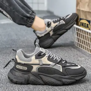 A person in dark joggers wears Factory Wholesale OEM ODM Summer Mesh Sneakers—black and gray chunky sports shoes with thick, anti-slip PU rubber soles—seen on a carpeted floor near an IKEA bag.
