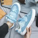 Wearing black jeans and a silver ring, a person tries on 2025 Wholesale ODM Custom Summer New Canvas Sneakers—light blue and white plaid lace-up shoes with thick white rubber soles.