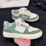 A hand holds a Factory Wholesale Retro Breathable Men Sneaker in green and white with cream laces; another matching sneaker is on a black surface in the background.