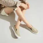 Person seated on a light surface, wearing light shorts, a plaid shirt with rolled sleeves, and Fashionable Casual Waterproof PU Upper Slip on Flats Shoes Elastic Band Flats Walking Shoes for Women in beige.