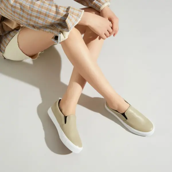 Person seated on a light surface, wearing light shorts, a plaid shirt with rolled sleeves, and Fashionable Casual Waterproof PU Upper Slip on Flats Shoes Elastic Band Flats Walking Shoes for Women in beige.