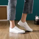 A person in frayed-hem blue jeans and Hot Sale Customize Fashionable PU Upper Women's Canvas Shoes stands on a wooden floor with decor in the background.
