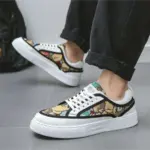 A person in black pants wearing Wholesale Factory 2024 Rubber Embossing Canvas Shoes—laced, multicolored with a leaf pattern, white soles and laces—stands on a gray floor.