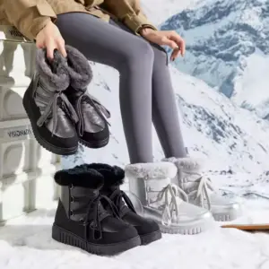 A person in gray leggings sits on a white bench in the snow, holding Fashion Leather Ladies Winter Snow Hiking Boots; black and silver pairs of these waterproof outdoor boots are placed on the ground nearby.