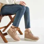 A person sits on a wooden chair, wearing blue jeans and Fashion Leather Low Heel Plus Size Winter Snow Hiking Boots for Men Women—lace-up, stylish boots—perfect for highlighting OEM/ODM fashion on a plain light background.