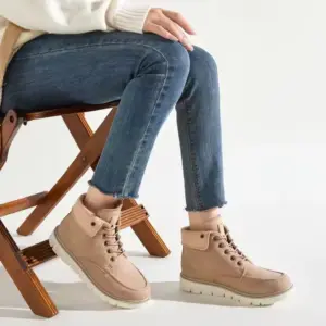 A person sits on a wooden chair, wearing blue jeans and Fashion Leather Low Heel Plus Size Winter Snow Hiking Boots for Men Women—lace-up, stylish boots—perfect for highlighting OEM/ODM fashion on a plain light background.