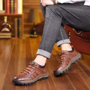 A person sits cross-legged in rolled-up gray jeans and Men's Solid Faux Leather Lace up Shoes Sneakers for Men—high-quality walking style shoes—on a wooden floor with books and a decanter, perfect for modern living spaces.