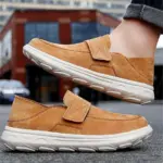 A person is wearing New Retro Men Shoes—light, shock-absorbing, flat leather loafers—in tan suede with thick white rubber soles, standing outdoors on a street with modern brick buildings in the background.