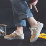 A person in cuffed blue jeans and tan 2025 Customized Wholesale Retro Street Trending Women Shoes Flat Sneakers stands on one foot, pointing at the lifted sneaker against a dark background.