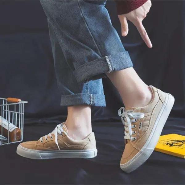 A person in cuffed blue jeans and tan 2025 Customized Wholesale Retro Street Trending Women Shoes Flat Sneakers stands on one foot, pointing at the lifted sneaker against a dark background.