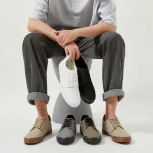 A person sits on a stool holding white and black XRH Factory New Customized Logo Men Canvas Sneakers, with three pairs of beige, gray, and tan lace-up casual walking flats for men displayed on the floor.