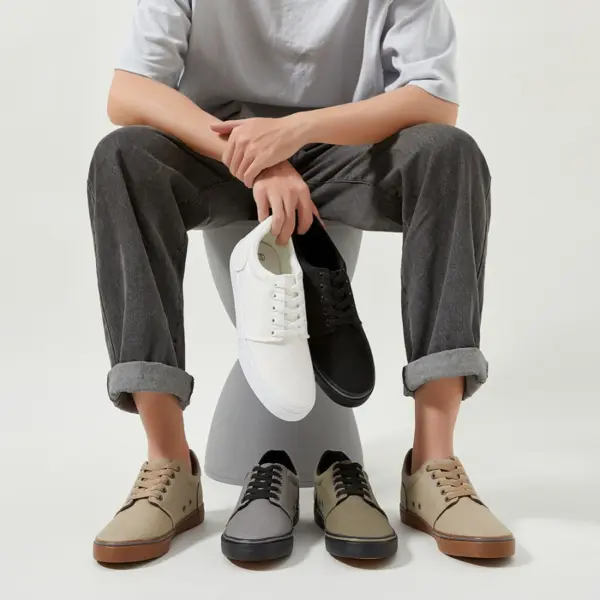 A person sits on a stool holding white and black XRH Factory New Customized Logo Men Canvas Sneakers, with three pairs of beige, gray, and tan lace-up casual walking flats for men displayed on the floor.