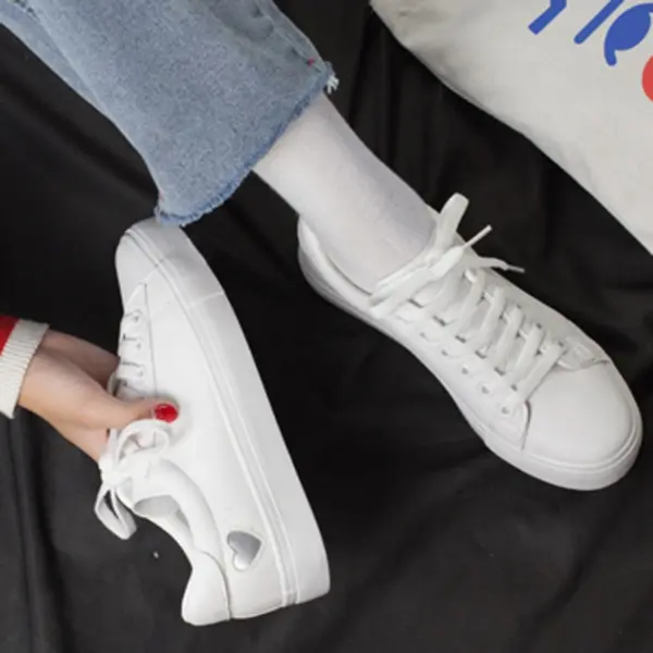 A person in white sneakers and blue jeans sits on a black surface, holding the Xrh Wholesale Spring PU Valentine's Day Gift Love Custom Shoes—low top vulcanized white shoes for women with a heart-shaped cutout.