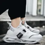 A person wears Wholesale Breathable Athletic Fitness Jogging Tennis Shoes Sport Running Sneaker for Men with transparent soles and a black tag, standing on concrete steps—ideal for OEM/ODM customization.