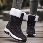 A person in OEM Factory High Top Breathable Warm Cotton Snow Boots—black, laced PU women’s shoes with white fur trim—paired with gray jeans stands on a wooden surface outdoors.
