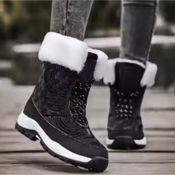 A person in OEM Factory High Top Breathable Warm Cotton Snow Boots—black, laced PU women’s shoes with white fur trim—paired with gray jeans stands on a wooden surface outdoors.