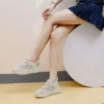A person in a blue denim skirt and 2025 Summer New Style Low Price Mesh Round Toe Sneakers, seated on a white circular object against a light background, highlights the fashionable versatility of these lace-up women’s shoes.