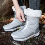 A person zipping up Wholesale Custom Winter Trendy Waterproof Snow Boots with plush lining stands outdoors on dry leaves and pine needles.
