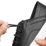 A person is threading a black elastic OEM ODM shoelace through the eyelets of Fashion Low Top PU Upper Casual Shoes—black flat walking canvas sneakers for women with white soles.
