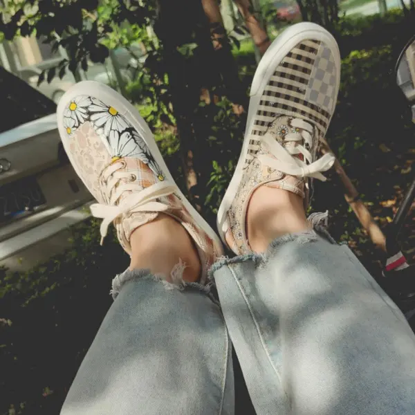 A person stands outdoors in light blue ripped jeans and mismatched OEM ODM Wholesale Mixed Wearable Canvas Shoes, with one sneaker featuring floral and daisy designs and the other boasting checkered paisley patterns.