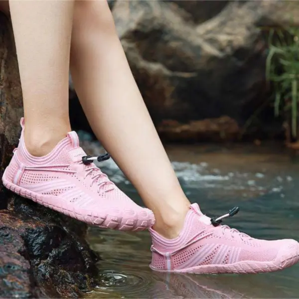 A person wearing light pink Customized Anti-slip Quick Drying Outdoor Shoes Flat Mesh Sports Flats Laced Beach Water Shoes sits by a rocky edge, dipping their feet into clear water outdoors.