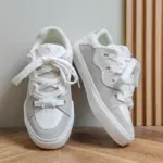 A pair of 2025 Wholesale ODM Custom Summer New Canvas Sneakers are on a wooden floor, one upright and the other on its side, showing their white and light grey lace-up design for women and men.