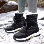 A person stands on snowy ground wearing grey pants and 2024 Customized LOGO Pattern Style Autumn Winter Trendi Warm Snow Boots Mixed PU Cotton Shoes in black and white with fur lining.