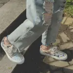 A person in ripped light blue jeans and Wholesale Mixed Wearable Canvas Shoes Lace up Breathable Sneakers Low Top Rubber slip on Loafers stands on a paved walkway and grass.
