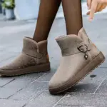 A person in black tights and taupe Wholesale Customized OEM ODM Plush Thick Waterproof Faux Fur Winter Boots Shoes Women Ankle Snow Boots stands on a gray tiled sidewalk.