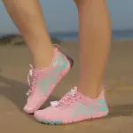 A person stands on sandy ground, one foot slightly raised, wearing Wholesale Customized Outdoor Beach Flat Anti-slip Breathable Sneakers Swimming Sports Children Shoes in pink and turquoise.