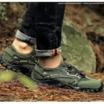 A person wearing green and black OEM ODM Wholesale Customized Outdoor Hollow Mesh Hiking Shoes with "Mashion" branding stands on a forest floor in rolled-up jeans, highlighting the shoes' anti-slip EVA rubber soles and breathable comfort.