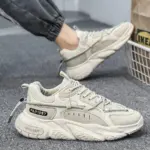 A pair of beige Factory Wholesale OEM ODM Summer Mesh Sneakers for men with decorative labels and stitching, shown on a gray carpet—one worn, one on display—highlighting customizable anti-slip outdoor PU rubber sports shoe options.