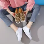 A person seated on the floor wears white slip-on sneakers and holds a pair of Hot Sale Casual Low Top No Lace Lazy Canvas Shoes Slip on Random Fluff Decoration Canvas Shoes for Women.