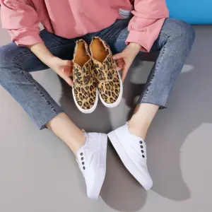 A person seated on the floor wears white slip-on sneakers and holds a pair of Hot Sale Casual Low Top No Lace Lazy Canvas Shoes Slip on Random Fluff Decoration Canvas Shoes for Women.