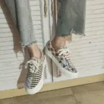 Wearing Wholesale Mixed Wearable Canvas Shoes—lace-up, breathable, low-top sneakers with floral, checkered, and plaid designs—this person sits by a white wooden cabinet, perfect for unique OEM ODM footwear needs.