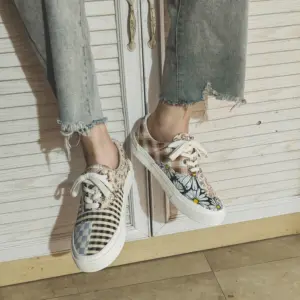 Wearing Wholesale Mixed Wearable Canvas Shoes—lace-up, breathable, low-top sneakers with floral, checkered, and plaid designs—this person sits by a white wooden cabinet, perfect for unique OEM ODM footwear needs.