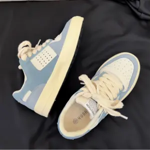 A pair of Factory Wholesale Retro Breathable Men Sneakers, featuring a cream and light blue design with thick cream laces, are displayed on a black fabric background.
