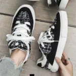 A person in distressed jeans tries on 2025 Wholesale ODM Custom Summer New Canvas Sneakers with lace-up design and solid rubber soles on a wooden floor.