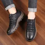 A person wearing Men's Solid Faux Leather Lace up Shoes Sneakers for Men High Quality Walking Style Shoes stands on a wooden floor, jeans rolled up at the ankles.