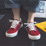 A person wearing 2025 Customized Wholesale Retro Street Trending Women Shoes—flat, solid color canvas sneakers with white laces and soles—stands on a dark floor in rolled-up blue jeans, with a yellow book partially visible nearby.