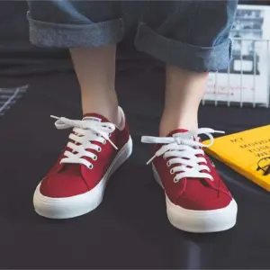 A person wearing 2025 Customized Wholesale Retro Street Trending Women Shoes—flat, solid color canvas sneakers with white laces and soles—stands on a dark floor in rolled-up blue jeans, with a yellow book partially visible nearby.