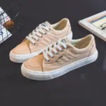 A pair of 2025 Customized Wholesale Retro Street Trending Women’s Solid Color Canvas Flat Sneakers in beige with white laces and rubber soles is displayed on a dark surface.