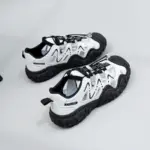 A pair of Factory Wholesale Hot Selling Thick Soled Sports Sneakers for women, featuring breathable mesh and PU, in white and black with metallic lace locks, displayed on a light gray surface.