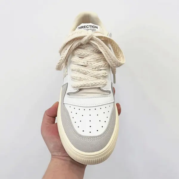 A hand holds a Factory Wholesale Retro Breathable Men Sneaker in white and beige with thick braided laces tied in a bow, viewed from above against a plain background.