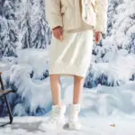 Wearing cream-colored quilted jacket, knit skirt, and Wholesale Customize logo Leather Sports Ankle Sneakers Boots for Women—snowproof and warm—she stands in snow with a snowy forest backdrop, showcasing ideal OEM ODM winter collection style.