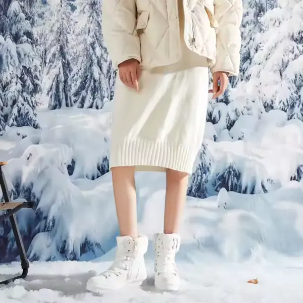 Wearing cream-colored quilted jacket, knit skirt, and Wholesale Customize logo Leather Sports Ankle Sneakers Boots for Women—snowproof and warm—she stands in snow with a snowy forest backdrop, showcasing ideal OEM ODM winter collection style.