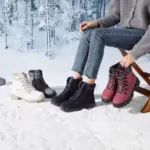 A person sits on a wooden chair in the snow, trying on four pairs of Wholesale Ankle Boots Snow Weather for Women and Men—leather, zipper, waterproof winter plush boots—in white, gray, black, and pink amid a snowy forest backdrop.